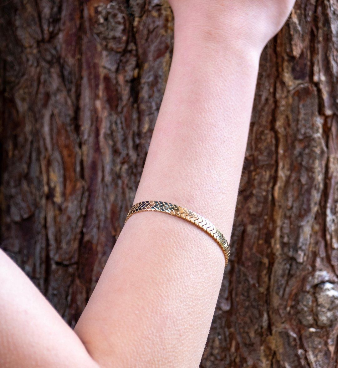 Slim herringbone-patterned gold bangle on an arm against a tree trunk