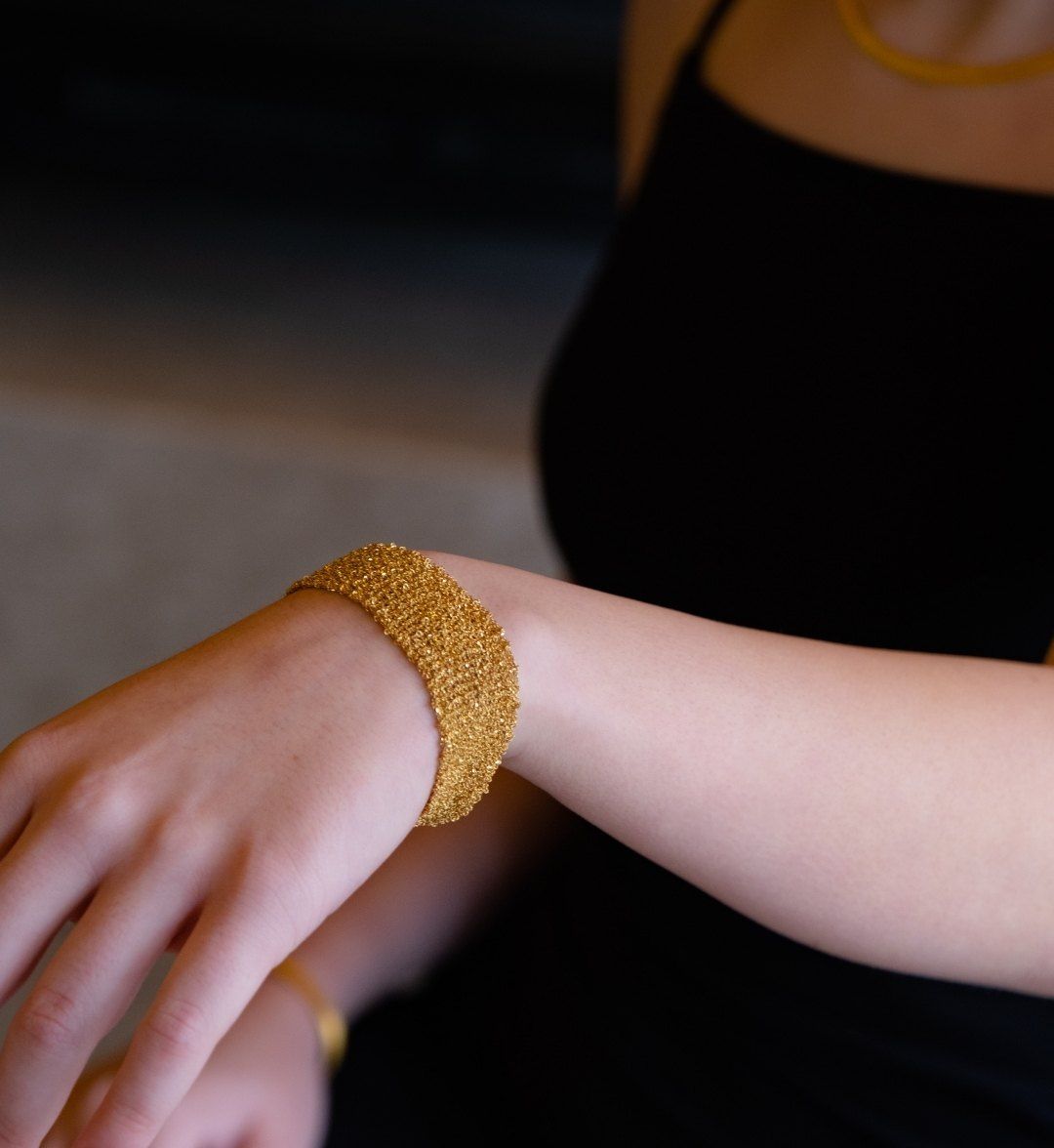 Wide woven yellow gold mesh bracelet on a model's wrist against a dark backdrop