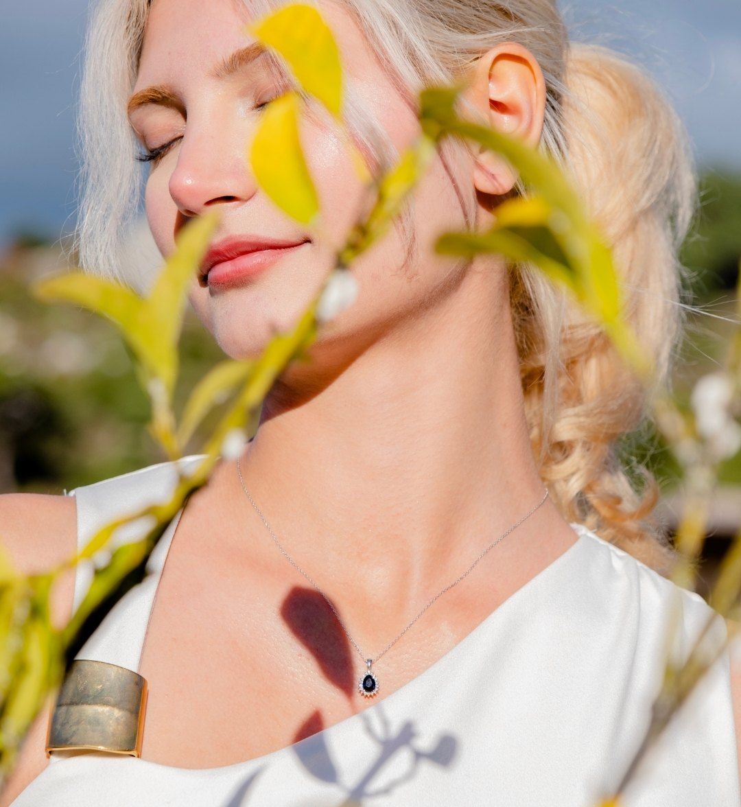 Navy teardrop pendant on a model among leaves, with a wide gold cuff on the arm