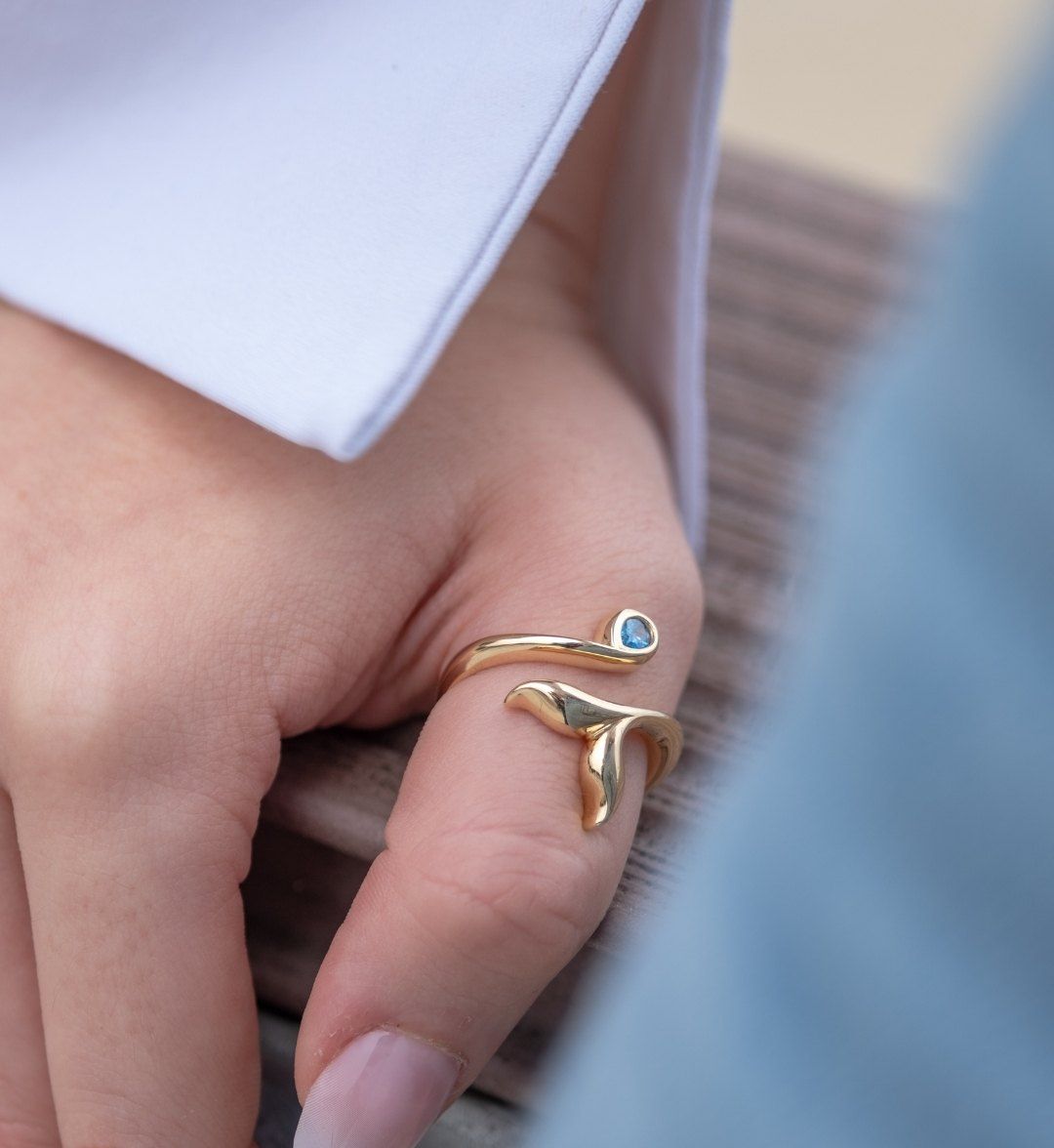 Open gold ring with a whale-tail motif and a blue stone at one end on a finger