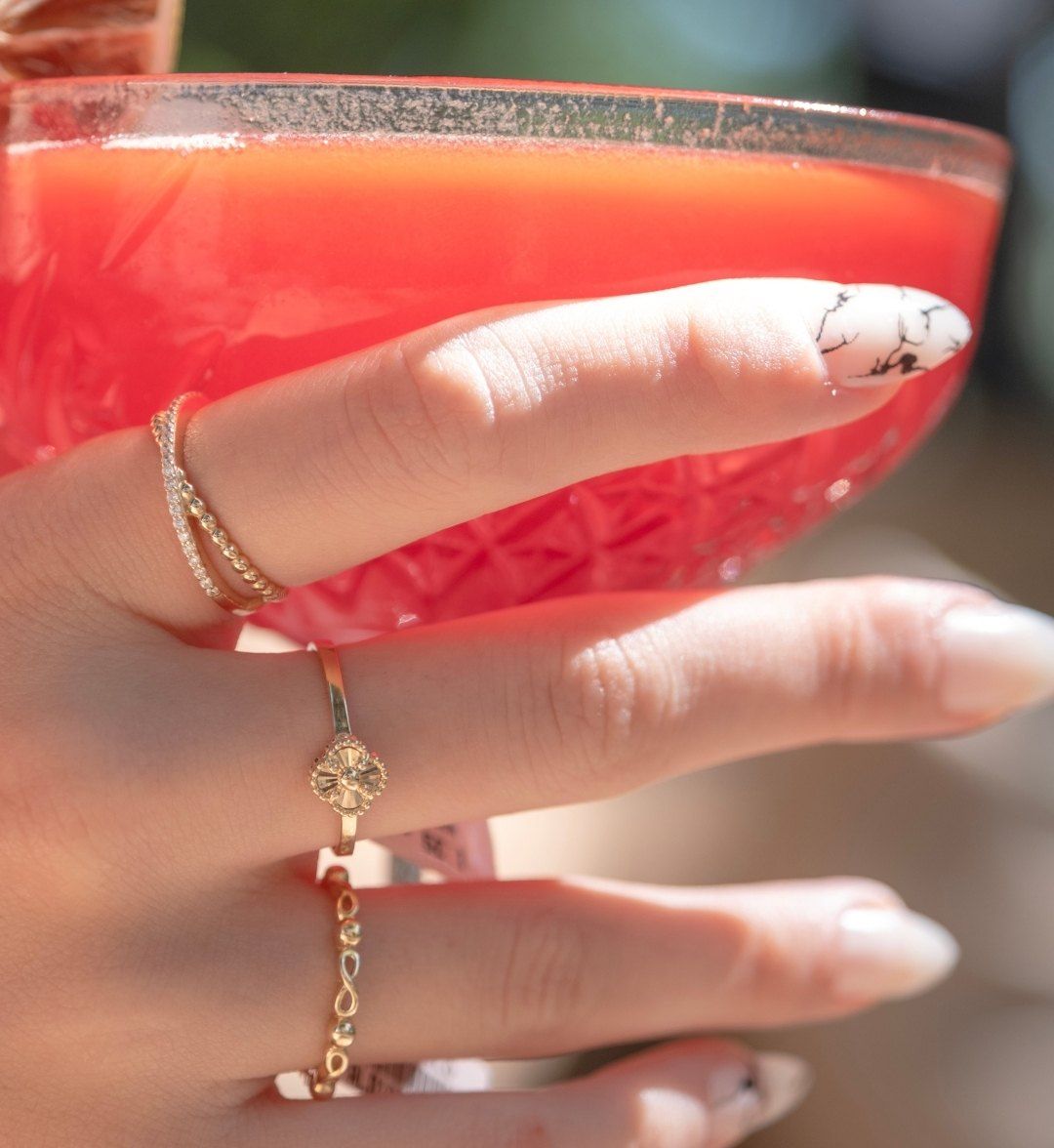 Three gold rings on a hand holding a glass: double-row stone, clover and infinity