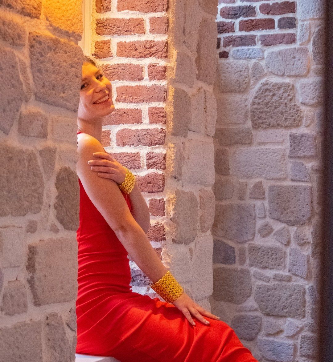 Model in a red dress by a stone wall, wearing wide woven gold cuffs on both wrists