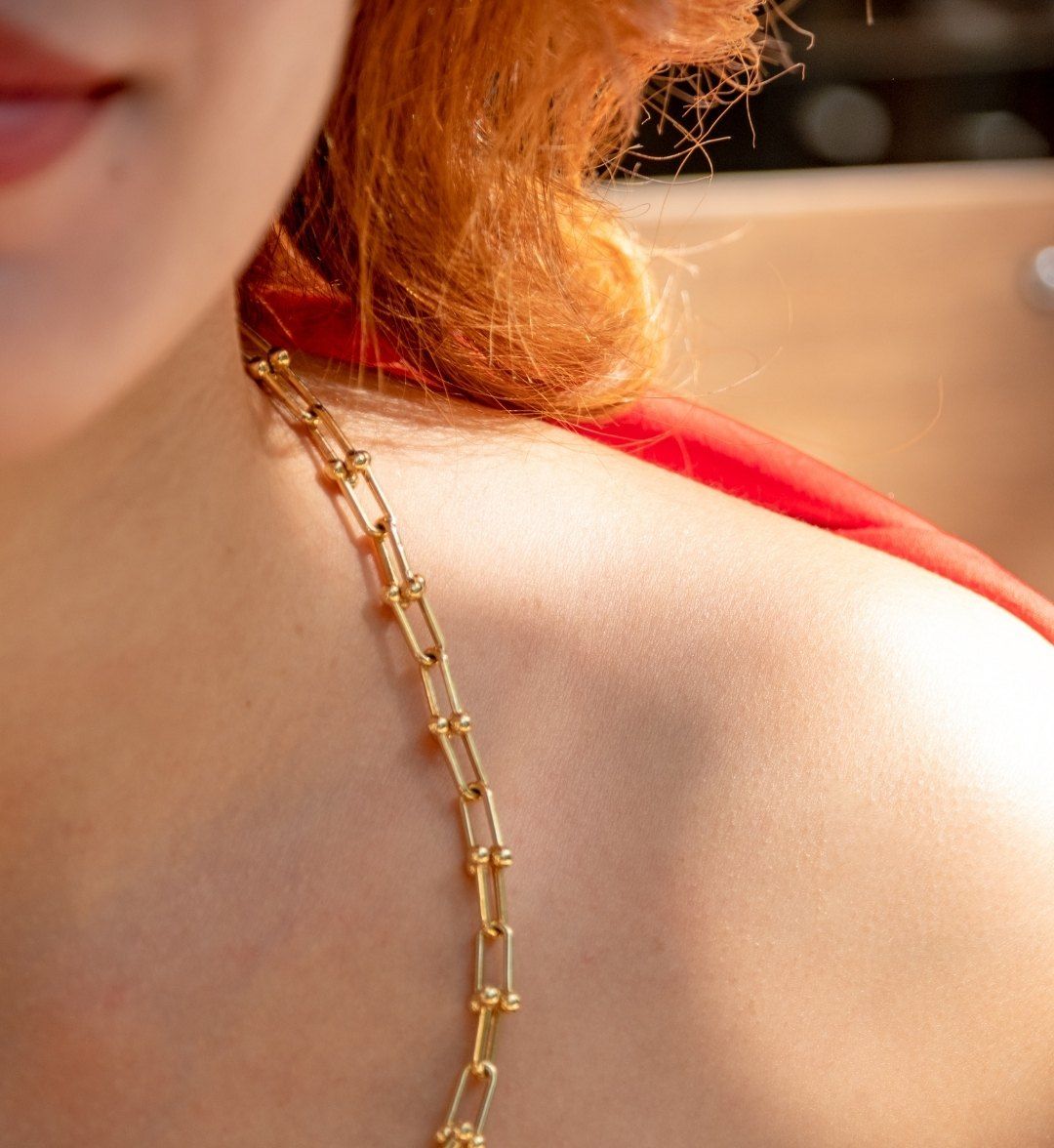Gold paperclip necklace with U-links and ball accents across a model's shoulder, red dress