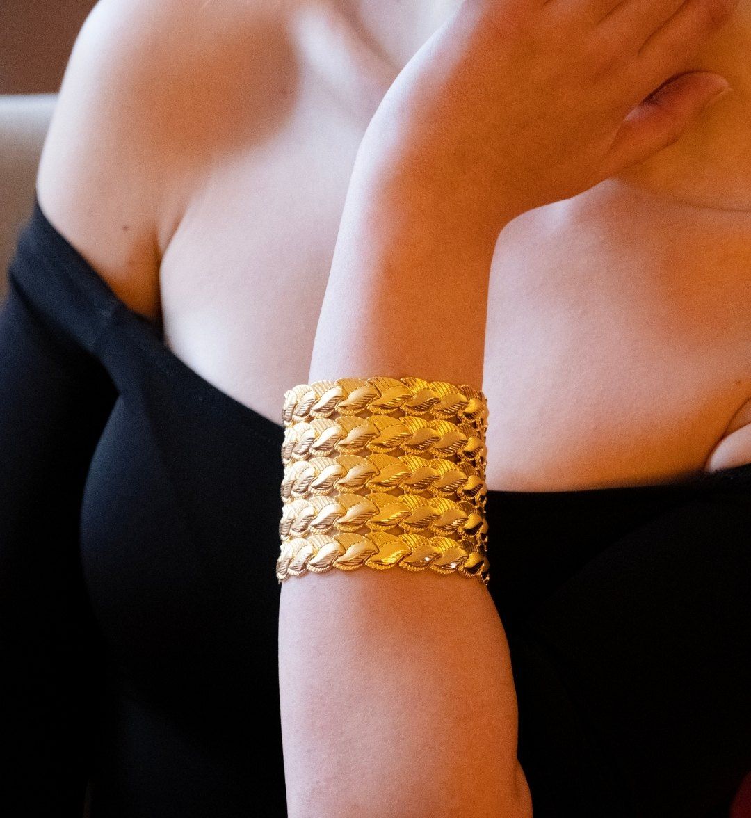 Wide woven gold cuff with five rows of leaf pattern on the arm of a model in a black dress