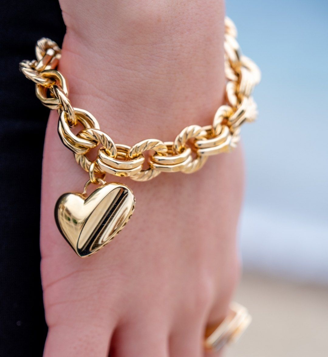 Puffed gold heart charm on a chunky twisted and plain link bracelet, close-up on the hand