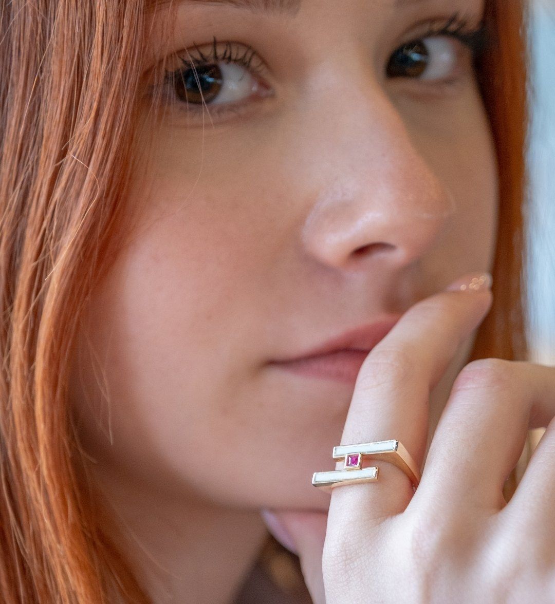 Open geometric gold ring with a pink stone on a model's finger, held near the face