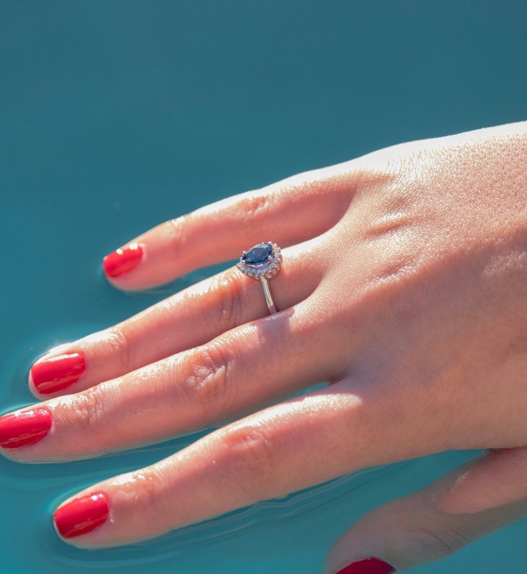 White gold ring with a navy pear-cut stone and white stone halo on a hand over water