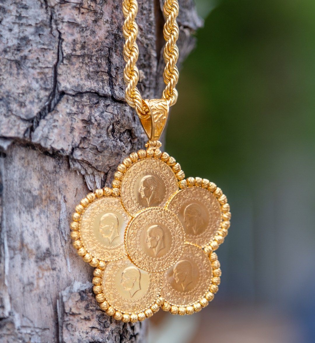 Flower-shaped pendant of seven gold coins on a rope chain, hanging against a tree trunk