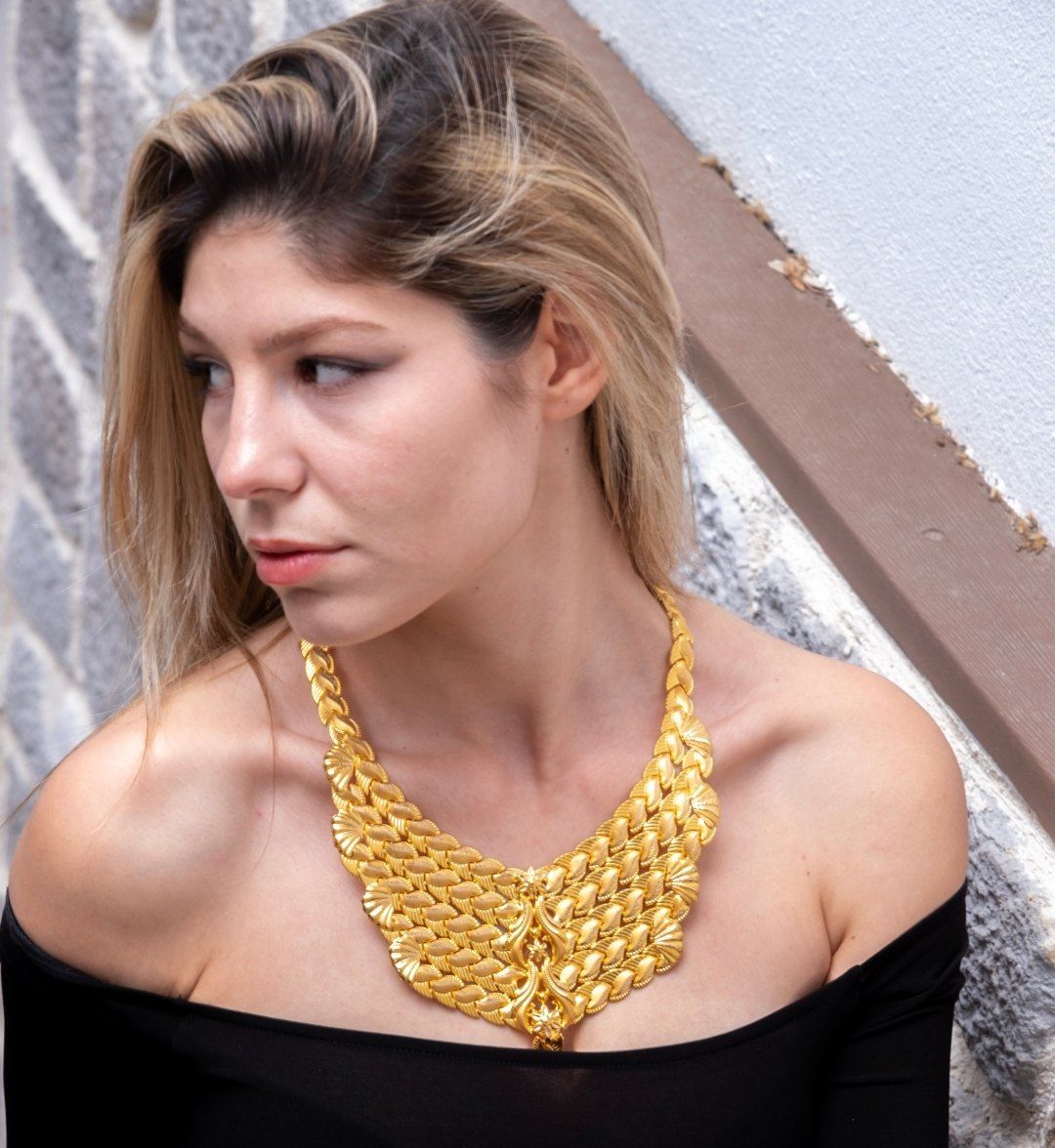 Wide layered gold collar with fan and leaf motifs worn by a model in an off-shoulder top