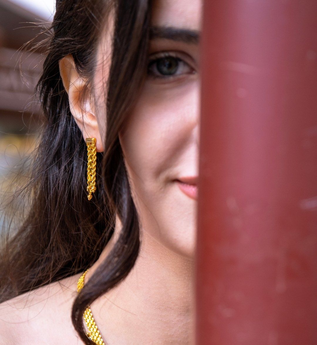 Long braided-chain gold tassel earring on a model, her face half hidden behind a post