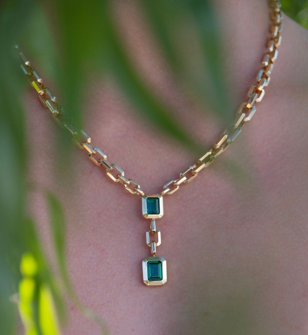 Green stone pendant necklace with two square stones on a chunky gold chain, seen through leaves